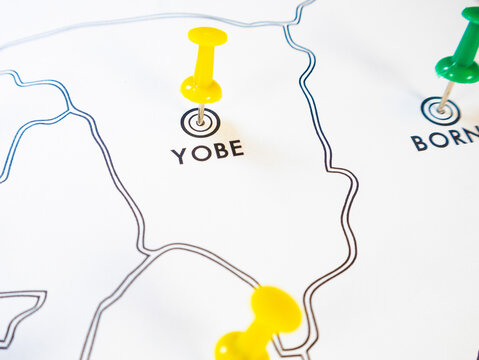 A close up shot of a white Nigerian state map. Brightly colored pushpins yellow and green mark specific northeastern regional areas including Yobe and Borno to pinpoint precise locations.
