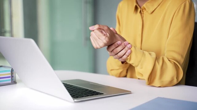 Close up of female hand holding and massaging painful wrist at office desk. Tired businesswoman suffering from carpal tunnel syndrome, joint inflammation and discomfort at modern workplace.