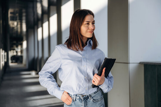 Young woman with brown hair wearing a light blue shirt and jeans stands confidently in a modern corridor, holding a tablet and smiling softly