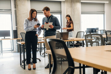 Fototapeta premium Two professionals discussing project details in a modern office space with desks and computers, while a woman works on a laptop in the background