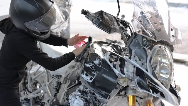 Close-up video of a woman in a black jumpsuit and helmet washes her motorcycle with a rag after applying foam at a self-service car wash. Woman takes care of her motorcycle. Slow motion 4k footage.
