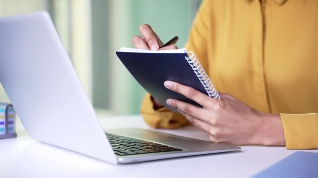 Close up of young businesswoman hands writing in notepad with pen. Professional female employee taking notes in notebook or planner during online business training or remote meeting at office.