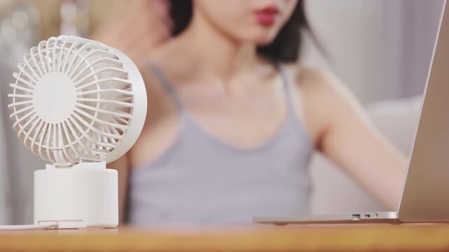 Tired woman sitting on sofa, working with laptop tablet in hot air struggle bad with lack of air condition, feels suffer from summer weather using portable handheld mini fan, cooling down, self-care