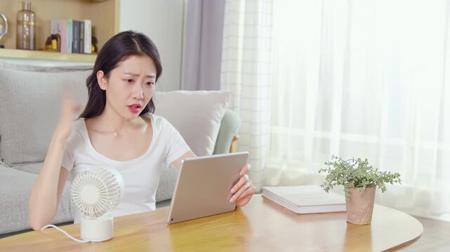 Exhausted young woman sitting, working in hot air, feels suffer from summer weather using portable handheld mini fan, tired lady sits on sofa with laptop tablet struggle bad with lack of air condition