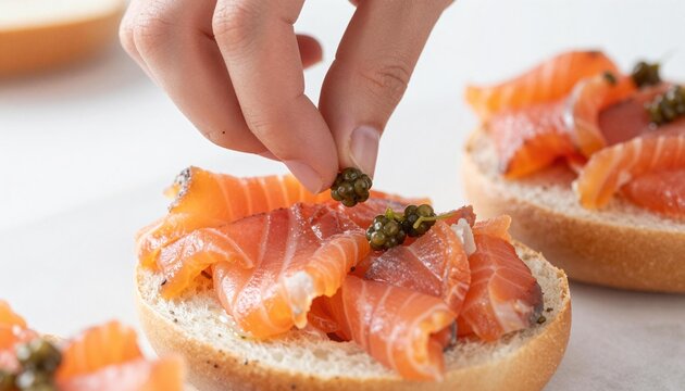 Hand placing a caper on a smoked salmon-topped toasted bagel bite