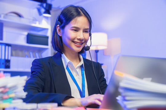 Smiling young Asian customer service representative wearing microphone headset and working on laptop in modern office.