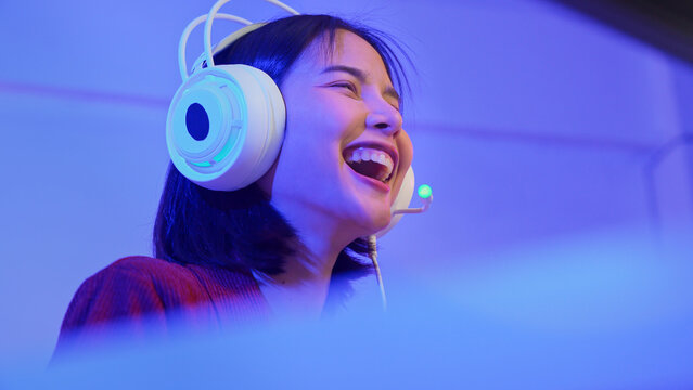 Excited young Asian girl playing video game with gaming headset and controller at home, esports online concept.