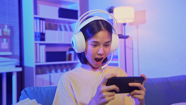 Excited young Asian girl playing mobile video game with headset and smartphone at home, online gaming and esports concept.