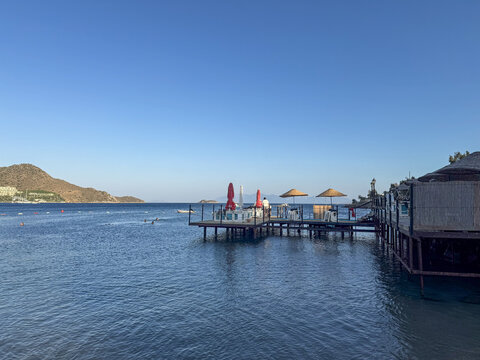 View of a wooden pier with red and straw umbrellas over the blue sea with swimmers and distant hills Bodrum, Turkiye.