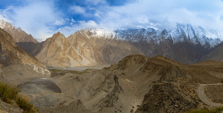 View of rugged snow-capped mountain peaks under a dramatic cloudy sky with arid rocky terrain and a winding road Gilgit-Baltistan, Pakistan.