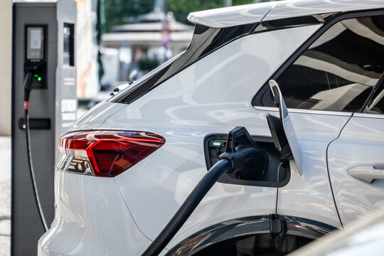 Nahaufnahme eines wei&szlig;en Elektroautos beim Aufladen an einer &ouml;ffentlichen Ladestation in einer st&auml;dtischen Umgebung bei Tageslicht