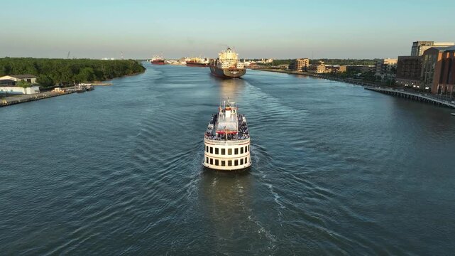 Aerial drone view of a riverboat cruise sailing down the Savannah River in Georgia, with a massive cargo ship and the downtown waterfront in the distance at golden hour.