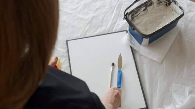 Woman arranges a sponge brush and palette knife on canvas at a covered work table at home. Concept of DIY textured wall art handmade decor and creative hobby.