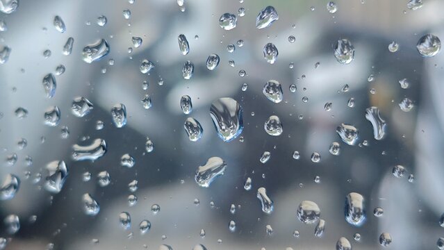 Clear water droplets on a transparent window glass pane surface with soft blurred background, creating a calm abstract texture during rainy day.
