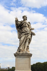 Fototapeta premium A detailed marble statue of an angel with flowing drapery set against a bright, partly cloudy sky, embodying classical baroque art.
