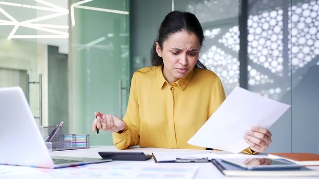 Confused young businesswoman having difficulty with paperwork and failing to understand report at office desk. Puzzled female employee looking stressed while working with complex business data.