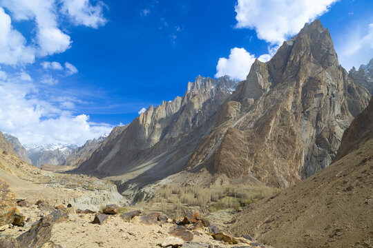 View of the towering granite peaks of the Karakoram range overlooking a remote mountain valley under a bright blue sky with scattered clouds in Gilgit-Baltistan, Pakistan.