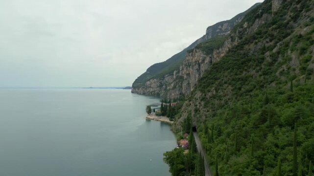 Aerial view gliding along forested cliffs above calm lake shoreline on cloudy day