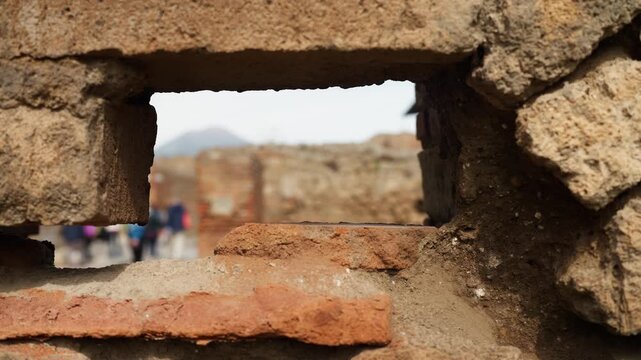 View through a small Pompeii wall opening with rough stone and brick in focus, framing blurred ruins and visitors beyond the ancient Roman site.