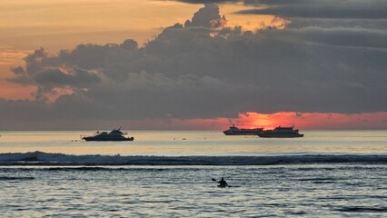 Sunrise view at Sanur Beach Bali featuring calm ocean waves, anchored boats, dramatic clouds, and warm orange sunlight reflecting across the sea. © mrbudiarto