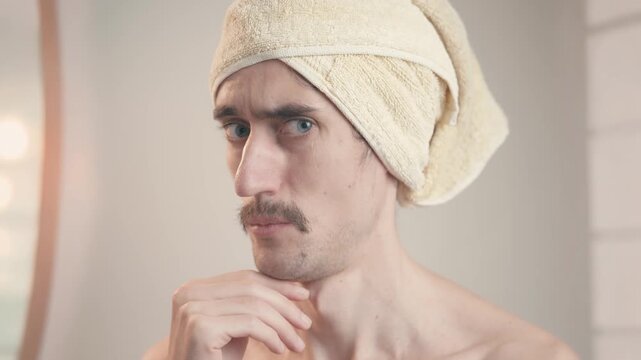 Comical mustached man in bathroom with towel on head looking at himself in mirror