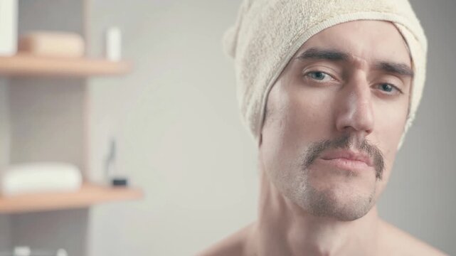 Funny mustached man in bathroom with towel on head staring seriously at camera