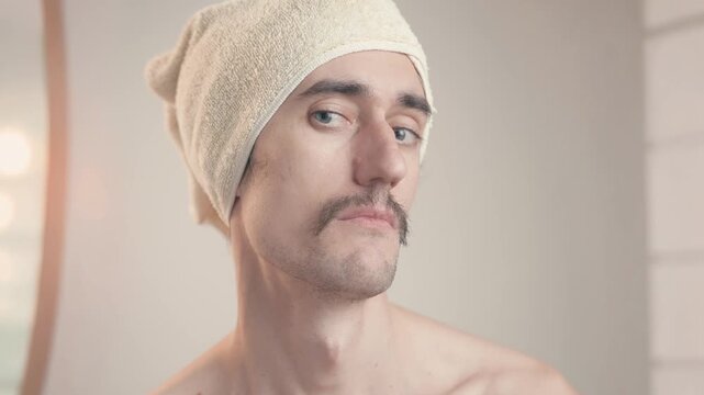 Mustached man in bathroom with towel on head sighs displeased, looking around in contemplation