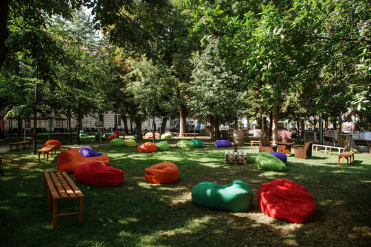 View of colorful bean bags and wooden benches scattered on a green lawn under the shade of lush trees in a public park Sremska Mitrovica, Vojvodina, Serbia.