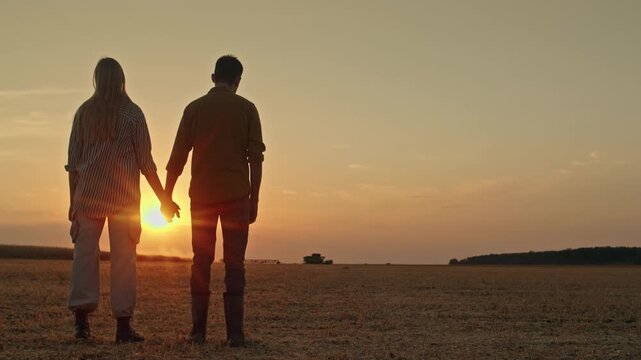 Farmers holding hands watching sunset over field with combine harvester in background footage.