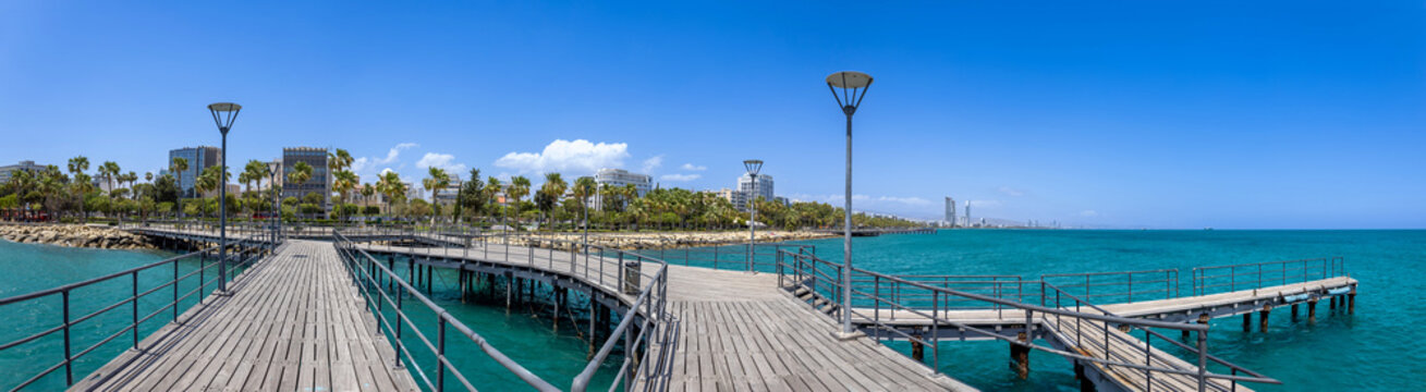 View of the wooden Molos promenade pier extending over the turquoise Mediterranean Sea with the city skyline and palm trees under a clear blue sky in Limassol, Limassol, Cyprus.