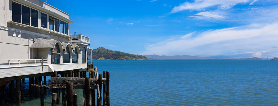 View of the Sausalito Boardwalk featuring a waterfront building on wooden pilings overlooking the blue waters of the bay and distant hills in Sausalito, California, United States.