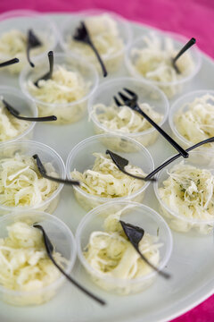 Clear plastic cups of shredded white cabbage salad with herbs, served with black forks on a white tray over a pink tablecloth