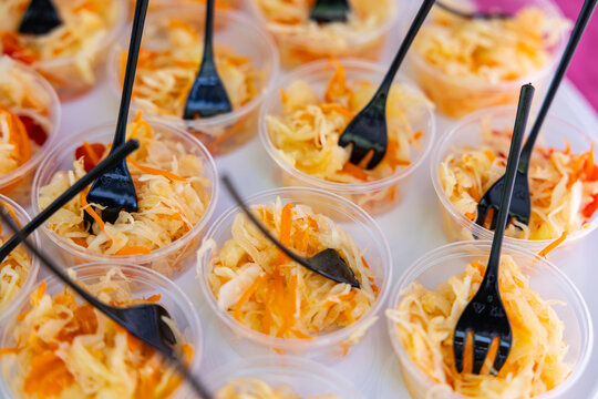 Clear plastic cups filled with shredded cabbage and carrot salad, each served with black forks on a white tray