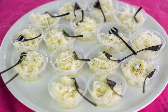 Clear plastic cups of shredded white cabbage salad with herbs, served with black forks on a white tray over a pink tablecloth