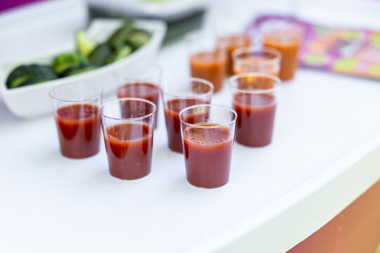 Small clear cups of tomato juice arranged on a white table with vegetables blurred in the background and copy space