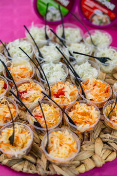Small plastic cups filled with shredded cabbage, carrot, and pepper salad, arranged on a woven tray with black forks on a pink tablecloth