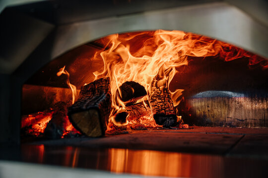 Intense flames and charred logs glowing inside a traditional wood-fired pizza oven, creating warm orange light and dramatic heat