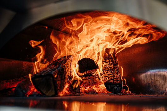 Intense flames and charred logs glowing inside a traditional wood-fired pizza oven, creating warm orange light and dramatic heat