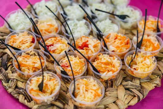 Small plastic cups filled with shredded cabbage, carrot, and pepper salad, arranged on a woven tray with black forks on a pink tablecloth