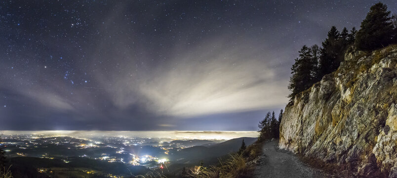View of the city lights from the Schockl mountain path under a starry night sky with rocky cliffs and pine trees overlooking Graz, Styria, Austria.