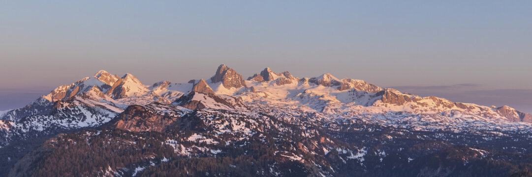 View of Dachstein glacier and the surrounding mountain peaks glowing in the warm light of sunset with snow-covered ridges and dense evergreen forests below Schladming, Styria, Austria.