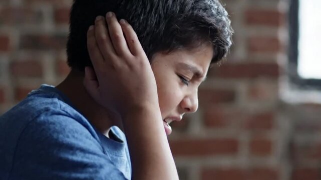 A young boy covering his ears in distress against a brick wall background