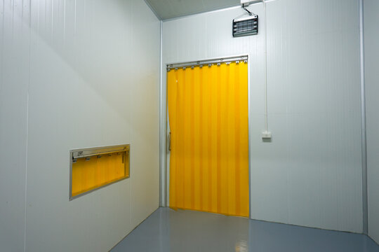 Empty industrial cold room interior featuring white sterile wall grey floor bright yellow plastic strip curtain door and small window conveying quiet emotion in modern factory warehouse