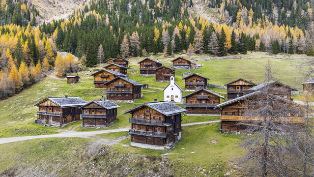 View of traditional wooden alpine huts and a small white chapel on a green hillside surrounded by colorful autumn larch trees Innervillgraten, Tyrol, Austria.
