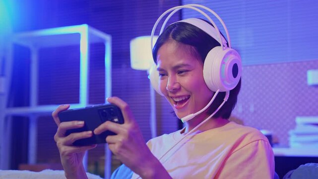 Excited young Asian girl playing mobile video game with headset and smartphone at home, online gaming and esports concept.