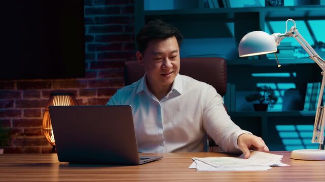 Asian business man opening laptop smiling working with internet and documents papers Korean businessman open computer device work paperwork online job smile at evening night office Chinese specialist