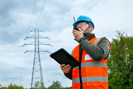 Worker using a portable radio for real-time reporting and damage prevention during a pre-storm site survey. Utility company technician or inspector evaluating electrical grid infrastructure. 