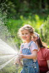 Happy mother and little daughter in denim overalls laughing and watering lawn with hose together.
