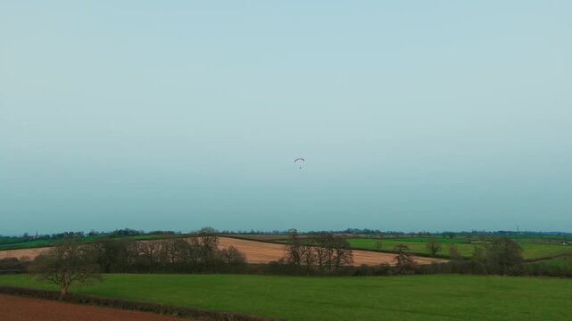 Distant paramotor pilot gliding over patchwork farmland fields forest edges and countryside panorama. Powered paragliding flight crossing agricultural landscape under calm sky extreme aerial