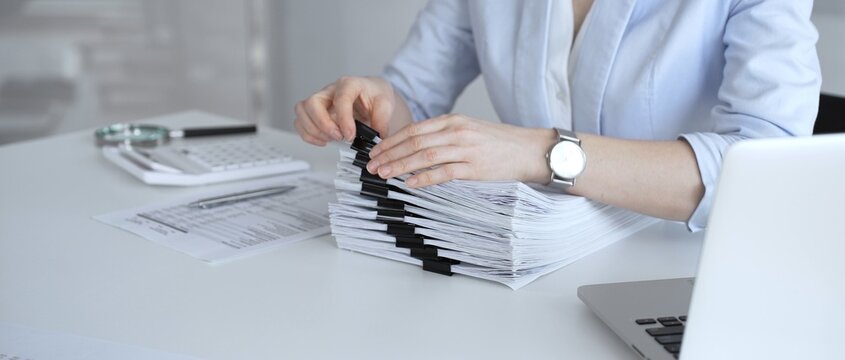 Businesswoman managing heavy workload, organizing financial documents for corporate accounting, tax preparation, and audit, working at office desk with stack of paper files and laptop. Audit and taxes